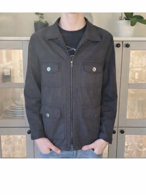 Svart jacka från Oscar Jacobson - Stilren svart jacka från Oscar Jacobson med fyra fickor framtill och dragkedja. Klassisk krage och raka ärmar. Jätte bra att använda till lite mer kyliga sommardagar. Skicket är i princip nytt, inget synligt slitage. Den är lite för stor för mig och knappast använd.