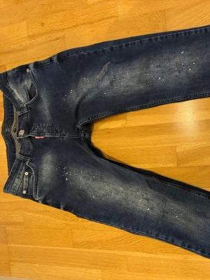 Mörkblå  jeans med slitningar - Säljer ett par mörkblå jeans från Dsquared2 med coola slitningar och vita färgstänk. Jeansen har klassisk femficksmodell, knappgylf och tydliga detaljer som ger en trendig och avslappnad look. Perfekta för dig som gillar streetstyle.
