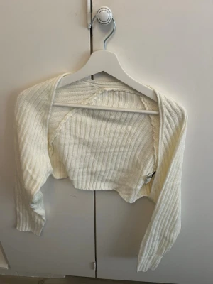 Vit cropped bolero i stickat material - Säljer en vit cropped bolero i ribbstickat material. Den har långa ärmar och är perfekt att slänga över ett linne eller klänning. Materialet är mjukt och stretchigt, vilket gör den bekväm att bära. Enkel och stilren design som passar till många outfits.