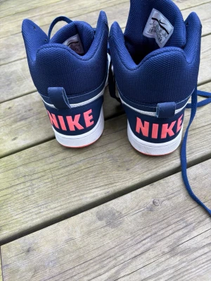 Blå Nike sneakers med vit swoosh - Snygga blå Nike sneakers med vit swoosh på sidorna och stora rosa NIKE-loggor på hälen. Skorna har högt skaft, blå snören och rosa yttersula. Ovandelen är i textil och syntetmaterial, perfekt för en sportig och trendig look.