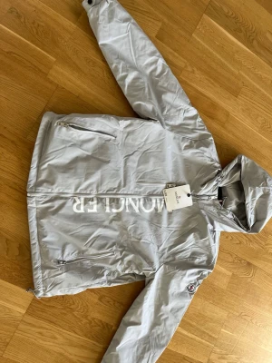 Grå vindjacka från Moncler - Snygg grå vindjacka från Moncler med huva och dragkedja framtill. Jackan har två snedställda fickor med dragkedja och en bröstficka. Stor Moncler-logga tryckt längs dragkedjan och klassisk patch på ärmen. Perfekt för blåsiga dagar.