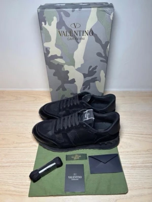 Svarta sneakers från Valentino Garavani - Snygga svarta sneakers från Valentino Garavani med diskret camomönster och detaljer i mocka och läder. Klassisk låg modell med svarta skosnören och logga på plösen. Perfekta för dig som gillar exklusiva och stilrena skor. Det har ett litet hål på sidan av skon skriv privat för att få se bild.