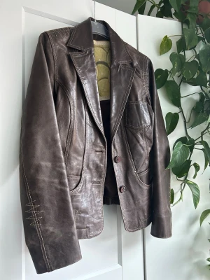 Brun skinnjacka med markerade sömmar - Snygg brun skinnjacka med klassisk krage och markerade vita sömmar. Jackan har två framfickor, knappstängning och dekorativa detaljer på ärm och rygg. Perfekt för dig som vill ha en stilren och tidlös look.