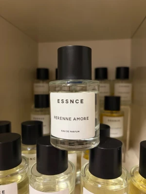 ESSNCE Perenne Amore Eau de Parfum - Fräsch parfym från ESSNCE med namnet Perenne Amore. Flaskan är genomskinlig med en minimalistisk vit etikett och svart rund kork. Parfymen har en stilren och modern design som passar perfekt på hyllan. Glasflaska med enkel och elegant look.