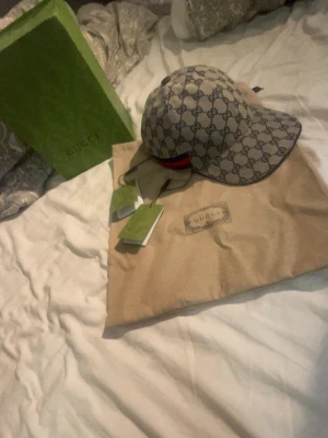 Gucci keps med monogram och röd detalj - Snygg keps från Gucci med klassiskt GG-monogram i grå och mörkblått. Kepsen har en böjd skärm och en röd-blå rand på sidan. Tillverkad i canvasmaterial och levereras med originalpåse och box.