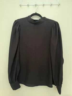 Svart blus med knytband i nacken - Stilren svart blus med djup v-ringning och knytband i nacken. Långa puffiga ärmar med breda manschetter och knappar. Tunt, lätt material som ger ett elegant fall. Perfekt för att lyfta din outfit med en trendig touch.