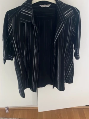 Svart randig skjorta med krage - Svart skjorta med vita ränder toch klassisk krage. Till dam. Skjortan har korta ärmar och en avslappnad passform, perfekt för en cool och stilren look. Materialet känns lätt och luftigt.