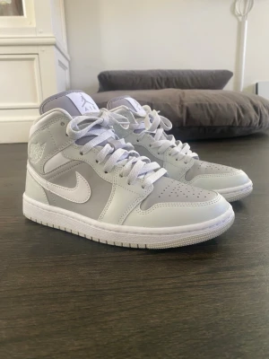 Nike Air Jordan 1 Mid grå/vit - Snygga Nike Air Jordan 1 Mid sneakers i grått och vitt med klassisk siluett. Skorna har vit swoosh, perforerad tå och högt skaft. Tillverkade i läder med textildetaljer och vita skosnören (använda en gång) 