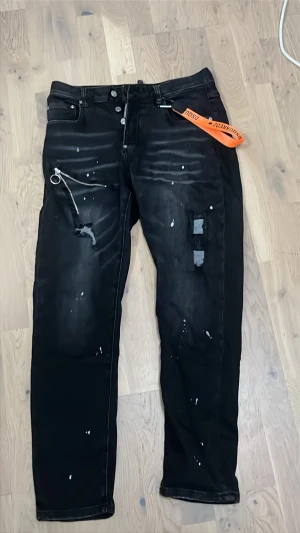 Svarta jeans med detaljer från Dsquared2 32/48 - Säljer ett par svarta jeans från Dsquared2 med coola slitningar, vita färgstänk och en orange tag i midjan. Jeansen har en smal passform, dragkedjedetalj och patch på bakfickan. Perfekta för dig som gillar streetstyle och unika plagg. Storlek 32/48. Från CNFans 