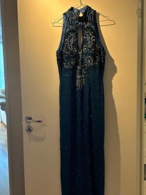 Mörkblå långklänning med paljetter - Supersnygg mörkblå långklänning med glittriga paljetter och broderade detaljer upptill. Klänningen är ärmlös med hög hals och nyckelhålsöppning både fram och bak. Perfekt för bal eller festkvällar när du vill glänsa extra.