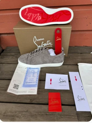  Louboutin skor loubs - Gråa sneakers i mocka från Christian Louboutin med klassisk röd sula och vita kanter. Lågt skaft och stilren design, perfekt för dig som gillar lyxiga detaljer. Skorna levereras med originalkartong, dustbag och kort. Ej autentisk.