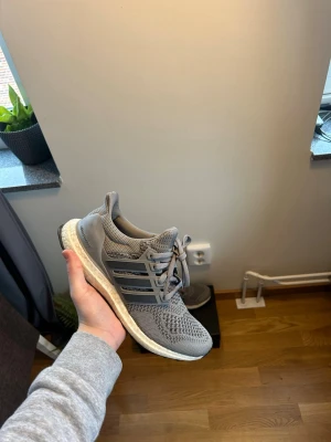 Grå Adidas Ultraboost sneakers - Säljer ett par grå Adidas Ultraboost sneakers med stickad ovandel och klassiska tre ränder på sidan. Skorna har en vit Boost-sula för extra komfort och gråa snören. Perfekta för dig som gillar stilrena och bekväma sneakers. Storlek 39 1/3