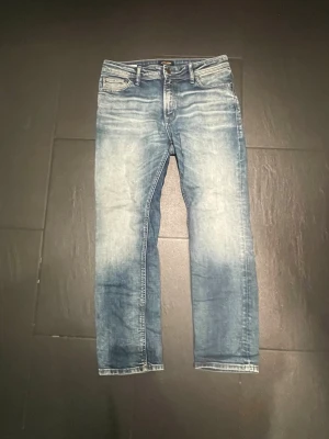 Blå faded jeans från Jack & Jones - Säljer ett par blå jeans från Jack & Jones med snyggt slitna detaljer och ljusa tvättade partier. Klassisk femficksmodell med normal midja och raka ben. Perfekta för en avslappnad och trendig look.