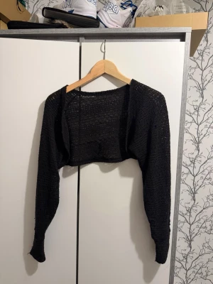 Svart stickad bolero - En svart stickad bolero med långärmad design och öppen front. Materialet är tunt och luftigt, perfekt att slänga över en topp eller klänning. Kort modell som slutar ovanför midjan och ger en cool layering-effekt.