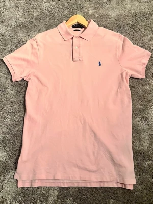 Ljusrosa pikétröja från Ralph Lauren - Säljer en ljusrosa pikétröja från Ralph Lauren med klassisk krage och knappar framtill. Tröjan har korta ärmar och är tillverkad i mjuk bomull. Den ikoniska blå loggan är broderad på bröstet. Perfekt för en clean och stilren look.