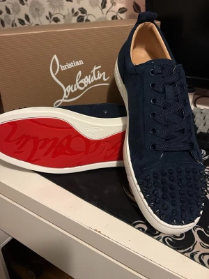 Blå Christian Louboutin sneakers - Snygga mörkblå sneakers från Christian Louboutin med klassiska röda sulor och coola nitar på tån. Skorna har en stilren design med snörning och är tillverkade i mocka. Perfekt för dig som vill sticka ut med exklusiva detaljer och premiumkänsla.