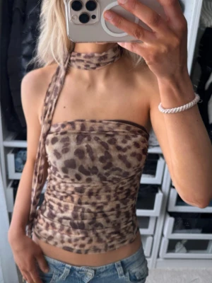 Leopard mönstrad tubtopp med scarf - Säljer en leopardmönstrad tubtopp i beige och bruna toner. Toppen är axelbandslös och har en matchande scarf som kan knytas runt halsen. Materialet känns stretchigt och lätt, perfekt för en trendig look. Från Zara. 