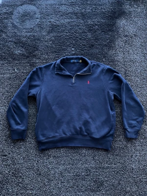 Ralph Lauren halfzip - Mörkblå sweatshirt med halv dragkedja och klassisk krage från Polo Ralph Lauren. Tröjan har en liten röd broderad logga på bröstet och ribbade muddar vid ärmslut och midja. Perfekt för en chill och stilren look. Storlek L, skick 9/10. Pris går att diskutera✅