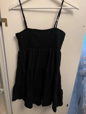 Svart kort klänning med smock - Svart klänning med smockad överdel och tunna axelband. Klänningen har en luftig och volangig nederdel som ger ett avslappnat och trendigt intryck. Perfekt för dig som gillar enkel och stilren design.