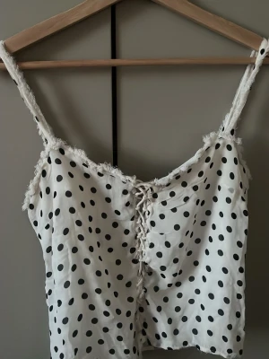 Prickig spetslinne från Zara Studio - Säljer ett vitt linne från Zara Studio med svarta prickar, tunna axelband och spetsdetaljer längs kanterna. Linnet har snörning framtill och är gjort i ett lätt, glansigt material som känns lyxigt och somrigt.