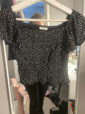 Svart prickig offshoulder topp KappAhl - Snygg svart offshoulder topp från KappAhl med vita prickar och puffiga korta ärmar. Smockad resår upptill och nertill ger en tight och bekväm passform. Perfekt att styla med jeans eller kjol för en trendig look.