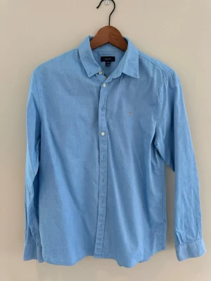 Blå skjorta från Gant i bomull - Klassisk ljusblå skjorta från Gant med broderad logga på bröstet. Skjortan har lång ärm, knappar framtill och är tillverkad i mjuk bomull. Perfekt till jeans eller chinos för en clean och stilren look.