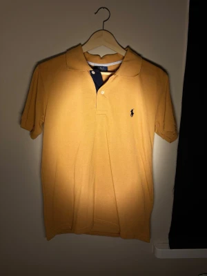 Gul pikétröja från Polo Ralph Lauren - Snygg gul pikétröja från Polo Ralph Lauren med klassisk krage och korta ärmar. Tröjan har en liten broderad logga på bröstet och knäppning framtill. Perfekt för en stilren och avslappnad look.