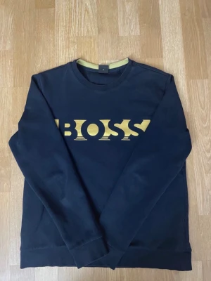 Svart sweatshirt från BOSS - Stilren svart sweatshirt från BOSS med guldfärgat logotryck på bröstet. Tröjan har rund halsringning och långa ärmar. Perfekt för dig som gillar en enkel men snygg look. Materialet känns mjukt och bekvämt.
