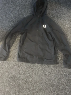 Svart vindjacka från Under Armour - Svart vindjacka från Under Armour med huva och dragkedja framtill. Jackan har två sidofickor och en vit logga på bröstet. Tillverkad i lätt och slitstark polyester, perfekt för blåsiga dagar.