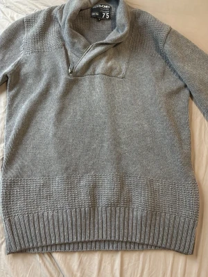 Grå stickad tröja Jack & Jones - Snygg grå stickad tröja från Jack & Jones med hög krage och dragkedja. Tröjan har ribbade muddar och ett diskret rutmönster upptill och nedtill. Perfekt för lager på lager och chill stil.