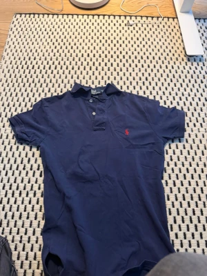 Mörkblå pikétröja från Ralph Lauren - Klassisk mörkblå pikétröja från Ralph Lauren med röd broderad logga på bröstet. Tröjan har korta ärmar, krage och knappar framtill. Perfekt för en stilren och avslappnad look.