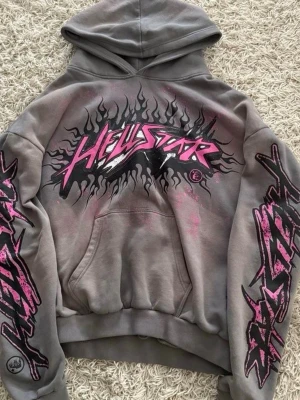 Grå Hellstar hoodie med rosa tryck - Säljer en grå hoodie från Hellstar med stor rosa och svart flames-grafik och logga på bröstet samt matchande tryck längs båda ärmarna. Hoodien har huva och magficka, och är gjord i mjukt bomullsmaterial. Perfekt för dig som gillar streetwear och statement-plagg.