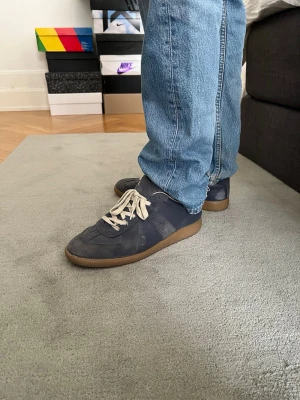 Mörkblå sneakers från Maison Margiela - Snygga mörkblå sneakers från Maison Margiela med klassisk design och beige gummisula. Skorna har vita snören och är tillverkade i en mix av mocka och läder. 