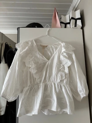 Vit volangblus med broderi - Söt vit blus med v-ringning, volanger och broderade detaljer framtill. Blusen har resår i midjan för en figurnära passform och långa ärmar med lätt puff. Blusen är helt ny i Stl S, nypris 299 men jag säljer den för 150 kr!