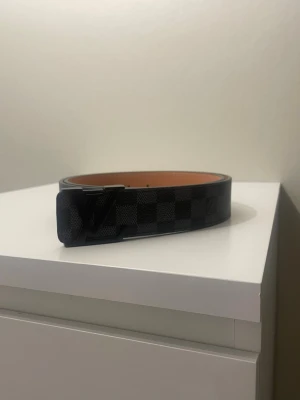 L V bälte  - Snyggt svart bälte från   med klassiskt rutigt mönster och -logga på spännet. Tillverkat i skinn med brun insida och präglad märkning. Perfekt accessoar för att lyfta din outfit.