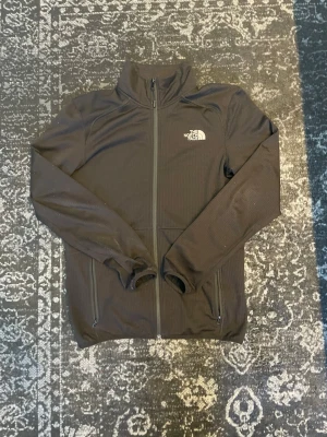 Svart ziptröja från The North Face - Svart ziptröja från The North Face med hög krage och dragkedja framtill. Tröjan har två fickor med dragkedja och logga på bröstet. Tillverkad i ett mjukt och stretchigt material som passar perfekt till en clean outfit