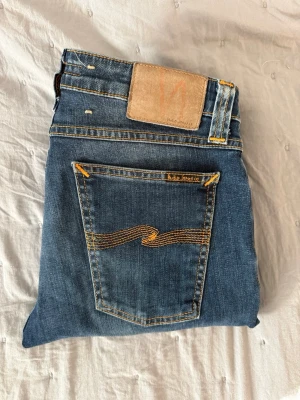 Blå jeans från Nudie Jeans - Säljer ett par klassiska blå jeans från Nudie Jeans med ikonisk gul vågsöm på bakfickan och läderpatch i midjan. Jeansen är i denim med snygga kontrastsömmar. Perfekta för dig som gillar stilrena och kvalitativa jeans.