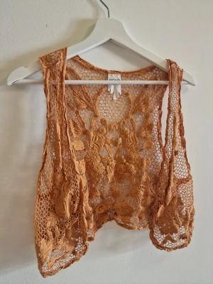 Orange spetsväst från Monki - Säljer en orange väst från Monki i spets med broderade blommor och öppen design. Perfekt att slänga över en topp för en cool och bohemisk vibe. Västen är tunn och har ett unikt mönster som sticker ut.