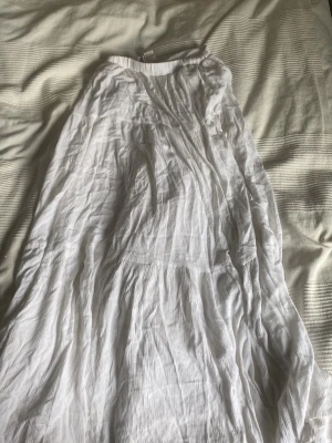 Vit långkjol i bomull - Luftig och flowy vit långkjol i mjuk bomull med resår i midjan. Kjolen har en lätt skrynklig struktur som ger en avslappnad vibe och passar perfekt till sommaren. Enkel och stilren design utan mönster eller extra detaljer.