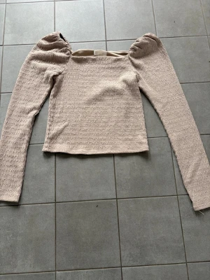Beige långärmad topp med puffärm - Trendig beige topp med långa puffärmar och fyrkantig ringning. Materialet är lätt strukturerat och stretchigt, vilket ger en snygg passform. Perfekt för dig som gillar en stilren men ändå unik look.