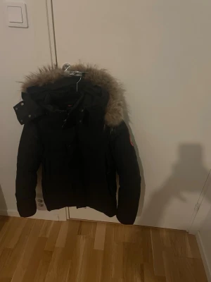 Svart jacka från Canada Goose - Säljer en svart jacka från Canada Goose med avtagbar huva och snygg pälskant. Jackan har klassisk logga på ärmen och stängs med dragkedja och knappar. Perfekt för kalla vinterdagar och riktigt varm.pris kan diskuteras 
