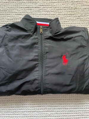 Svart vårjacka från Polo Ralph Lauren - Snygg svart vårjacka från Polo Ralph Lauren med röd broderad logga på bröstet och stor vit/röd logotyp på ryggen. Jackan har dragkedja framtill och står ut med sin sportiga look. Tillverkad i lätt, vindtätt material och har en hög krage med randig detalj.                                                                                               Äktenhet kan inte garanteras!