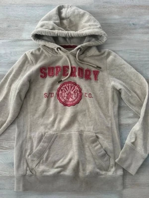 Beige hoodie från Superdry - Superfin hoodie i toppskick! Endast en deffekt som är en liten missfärgning som knappt syns väldigt inzoomad på bild 5. Skriv till mig vid eventuella frågor osv!