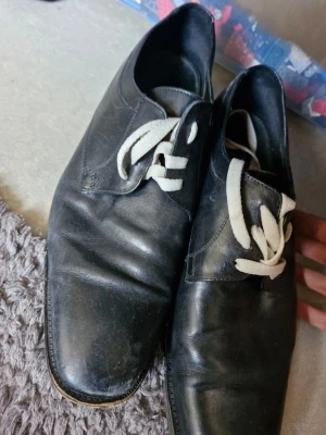 Svarta loafers med vita snören - Svarta loafers med klassisk design och vita snören. Skorna har en stilren form och är tillverkade i skinn, vilket ger en tidlös look. Perfekta för dig som vill ha en enkel men snygg sko till vardagsstilen.