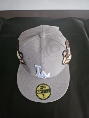 Grå LA New Era 59FIFTY keps - Snygg grå keps från New Era 59FIFTY med LA-logga framtill i vitt och broderade änglamotiv på sidorna. Klassisk flat brim och strukturerad passform. Perfekt för dig som vill sticka ut med coola detaljer och streetstyle-vibe.