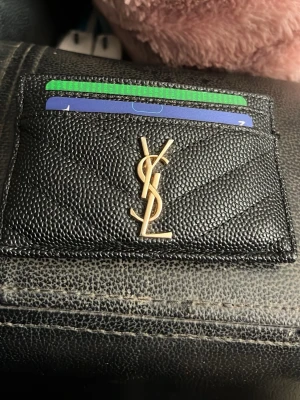 Svart YSL korthållare i läder - Stilren svart korthållare från YSL i präglat läder med guldfärgad logga framtill. Plats för flera kort och smidig att ha i fickan. Perfekt accessoar för dig som gillar lyxiga detaljer och klassisk design.