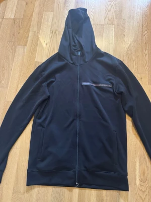 Svart hoodie från Prada med dragkedja - Svart hoodie från Prada med hel dragkedja framtill och huva. Diskret Prada-logga på bröstet i svart och rött. Tillverkad i ett mjukt och följsamt material som känns lyxigt. Perfekt för en stilren och avslappnad look.