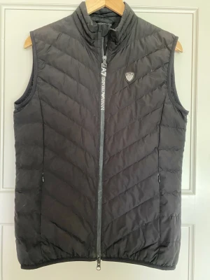 Svart dunväst från Emporio Armani - Snygg svart dunväst från Emporio Armani EA7 med hög krage och dragkedja framtill. Västen har ett stilrent EA7-märke på bröstet och diskreta sidofickor med dragkedja. Perfekt för lager på lager och streetwear-vibe. Knappt använd