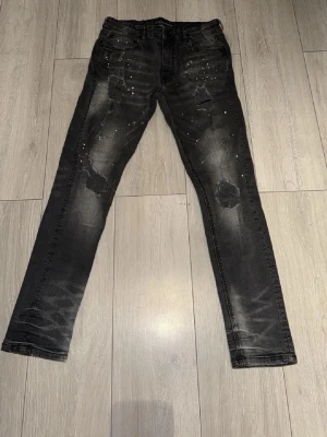 Svarta skinny jeans med slitningar - Säljer ett par svarta skinny jeans med slitningar och färgstänk på framsidan. Jeansen har en tvättad look, fem fickor och smal passform. Perfekta för dig som gillar en edgy och trendig stil.