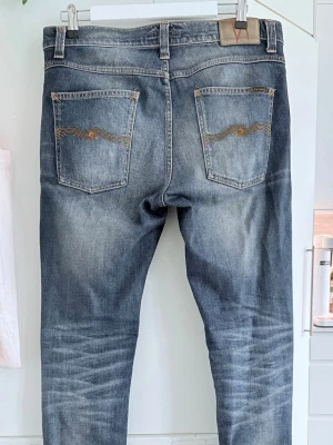 Blå Nudie Jeans - Säljer ett par blå jeans från Nudie Jeans med klassisk femficksmodell och snygga slitningar. Jeansen har kontrastsömmar och broderade bakfickor med Nudie-signaturen. Perfekta för en avslappnad och trendig look.